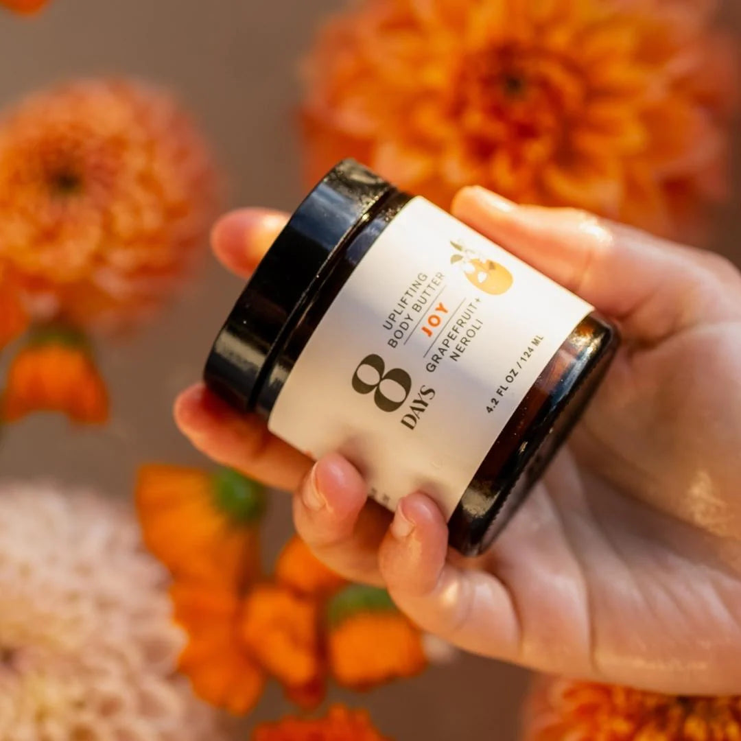A hand holding a jar of 8 Days Botanicals Organic Body Butter Joy over a field of orange flowers.