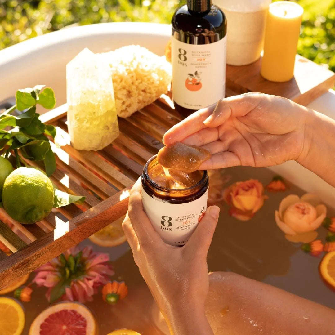 A bath shelf with 8 Days Botanicals Organic Body Polish - Joy being applied to a woman's hand.