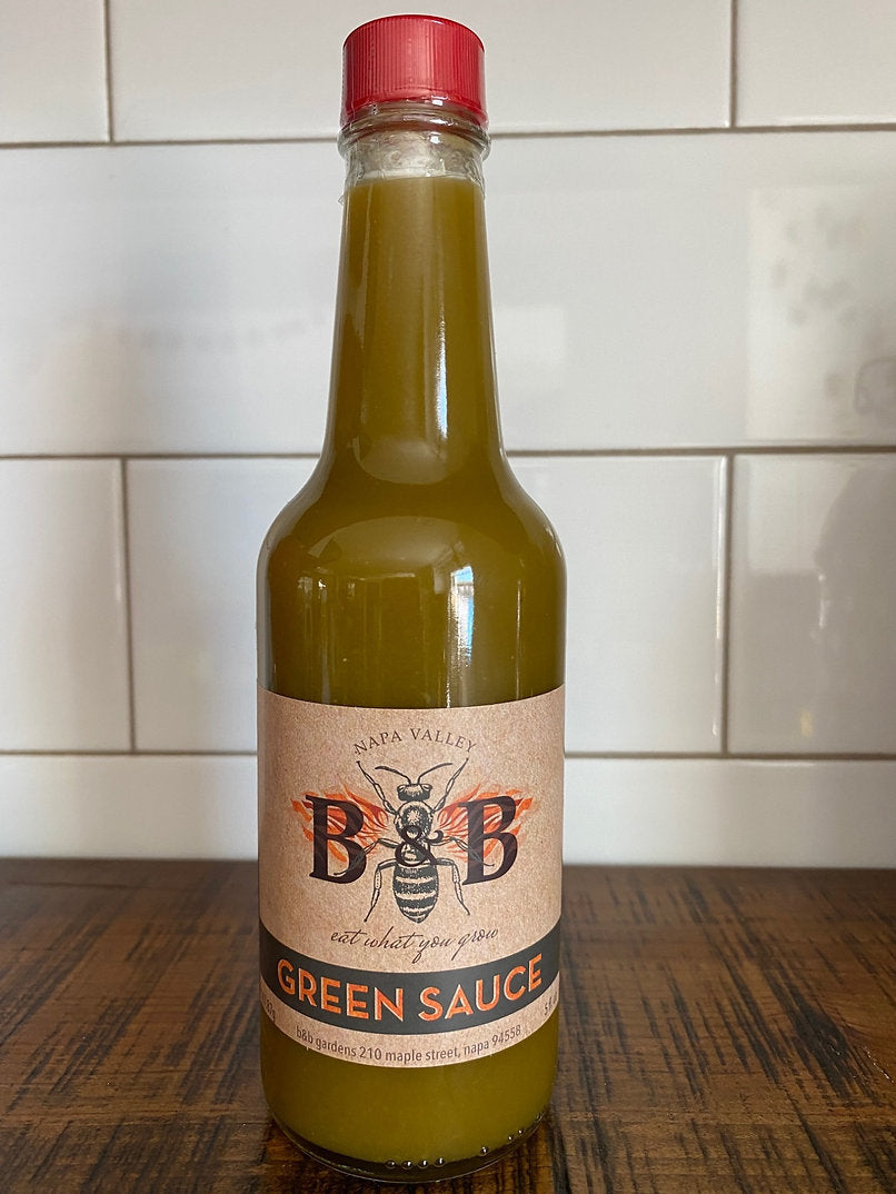 A greed bottle of B&B Gardens Hot Sauce - Green Sauce on a wood table against a white tiled wall.