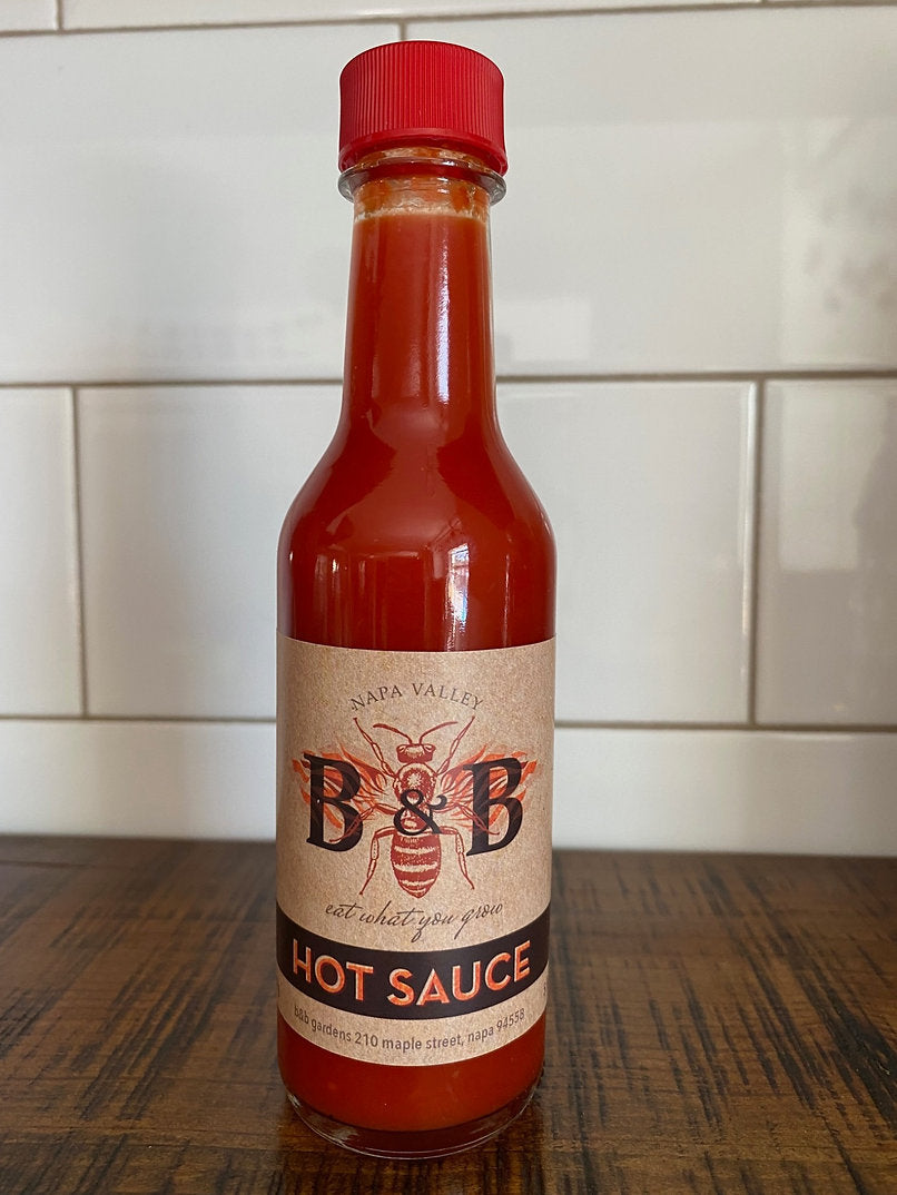 A red bottle of B&B Gardens - Hot Sauce on a wood table against a white tiled wall.