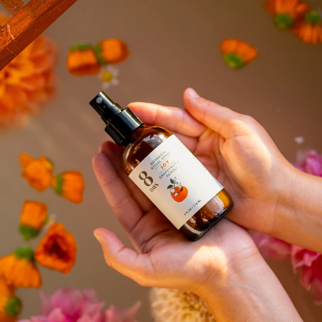 A person holding a spray bottle of 8 Days Botanicals Organic Room and Body Spray - Joy over a milky bath holding various orange and pink flowers.