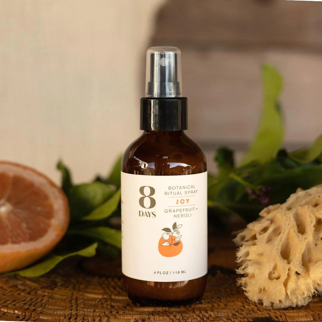 A spray bottle of 8 Days Botanicals Organic Room and Body Spray - Joy on a table next to a dried sponge and sliced grapefruit.