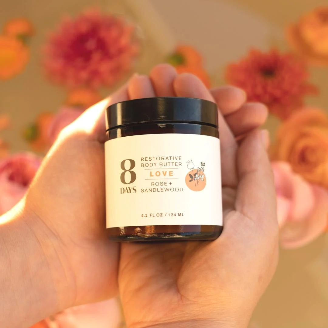 A person holding a jar of 8 Days Botanicals body butter over orange and red flowers.