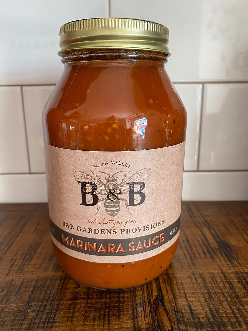 A jar of B&B Gardens Organic Marinara Sauce on a wood table against a white tiled wall.