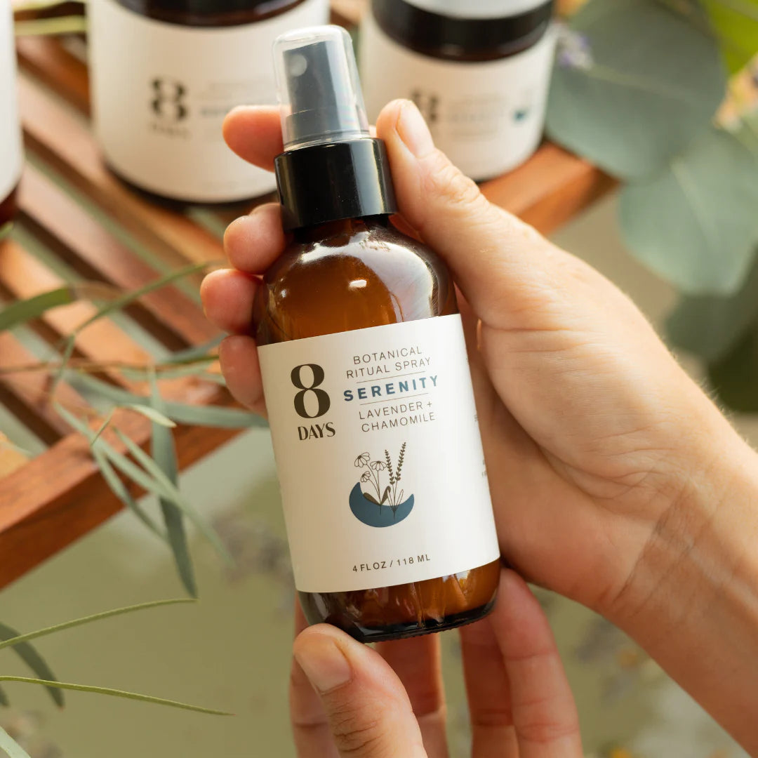 A person holding a bottle of 8 Days Botanicals Organic Room and Body Spray - Serenity over a milky green bath.