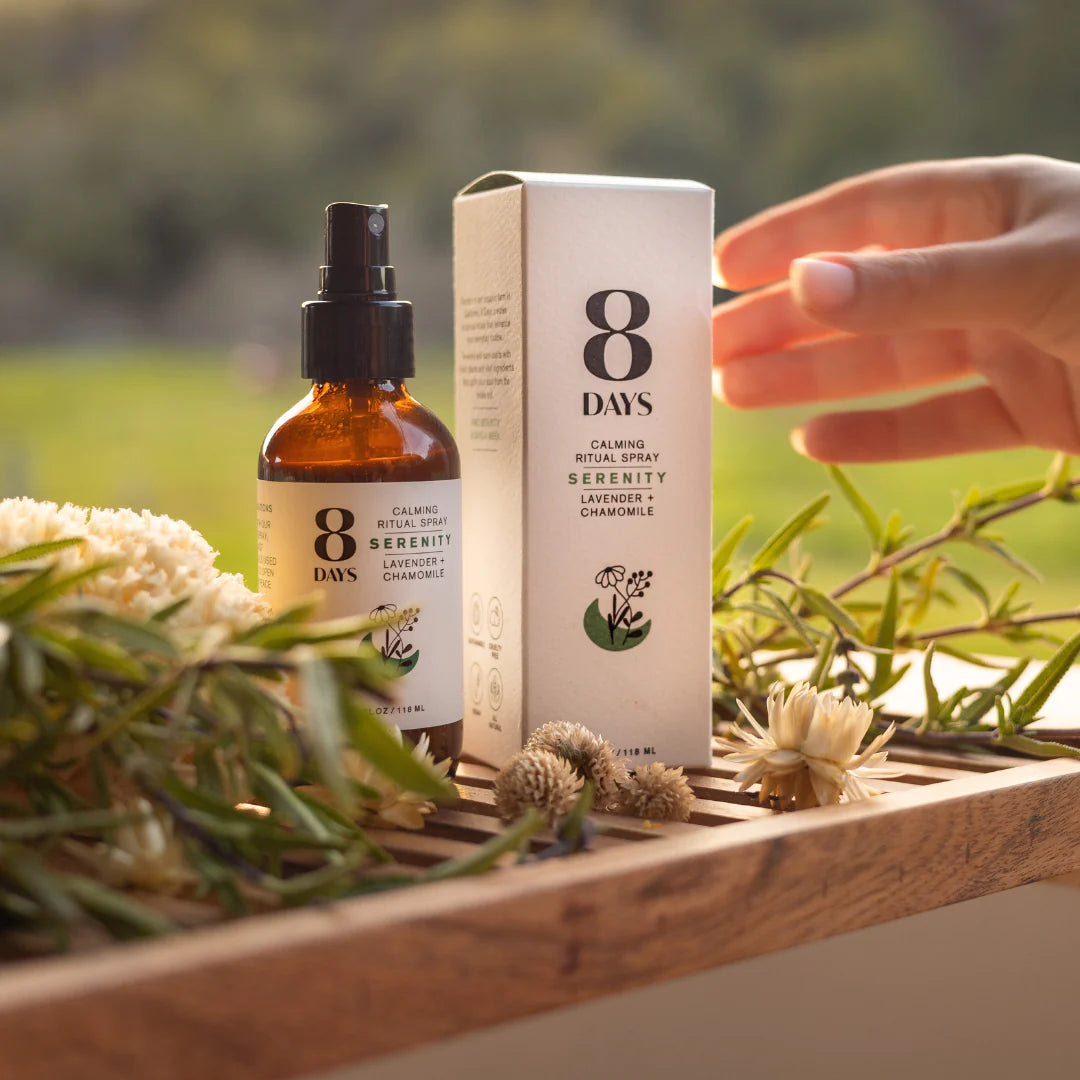 A person reaching to grab a bottle of 8 Days Botanicals Organic Room and Body Spray - Serenity.