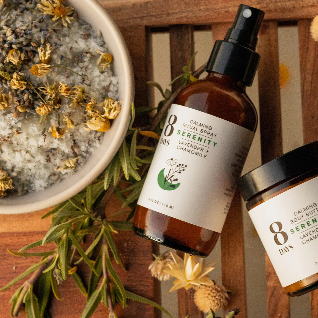 A bowl of chamomile, a twig of lavender and both the body butter and the spray bottle version of 8 Days Botanicals Organic Room and Body Spray - Serenity rest on a slotted table.