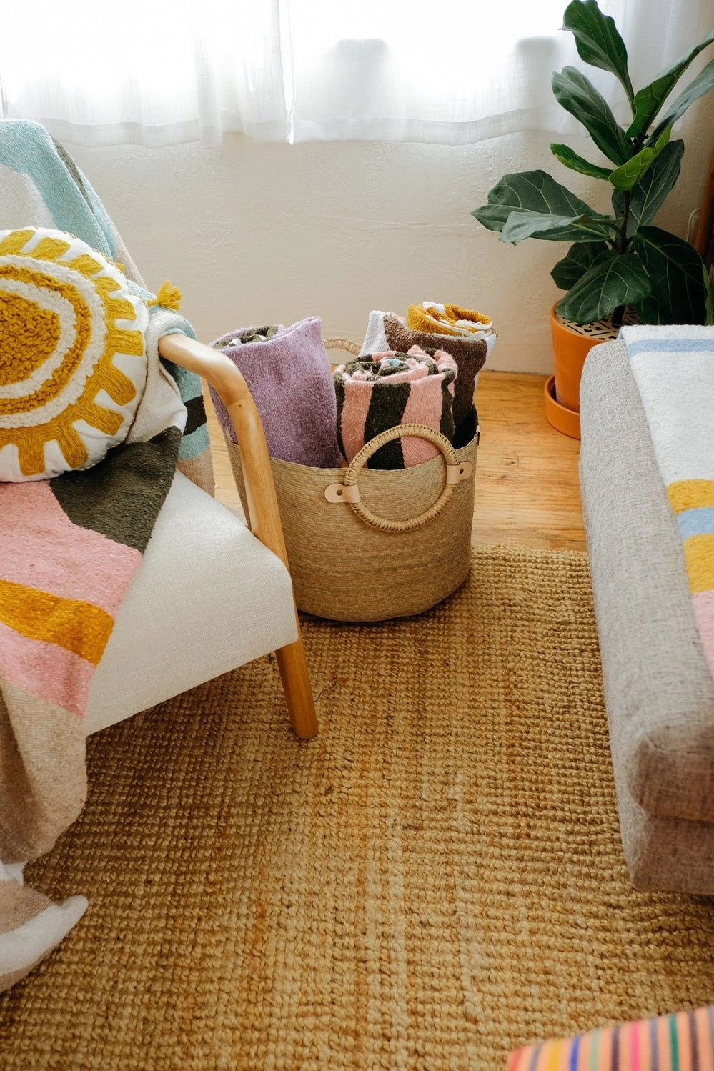 A Caminito Large Woven Floor Basket filled with colorful beach towels rests next to a chair in a sun room.