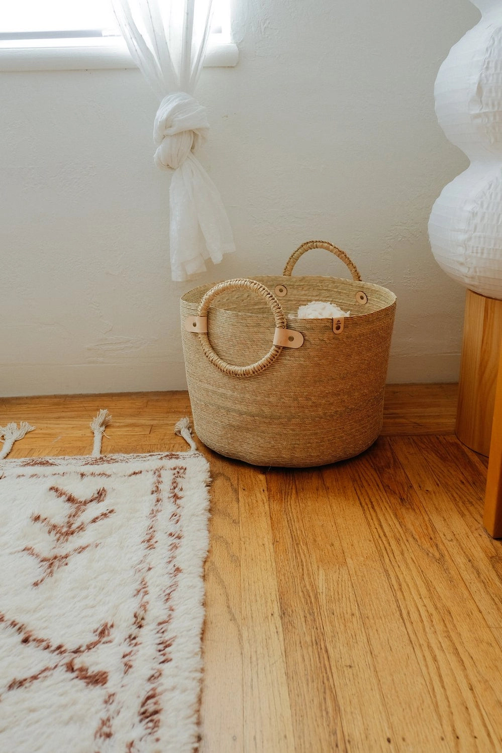 A Caminito Large Woven Floor Basket sits on a wooden floor next to a brown and white patterned rug.